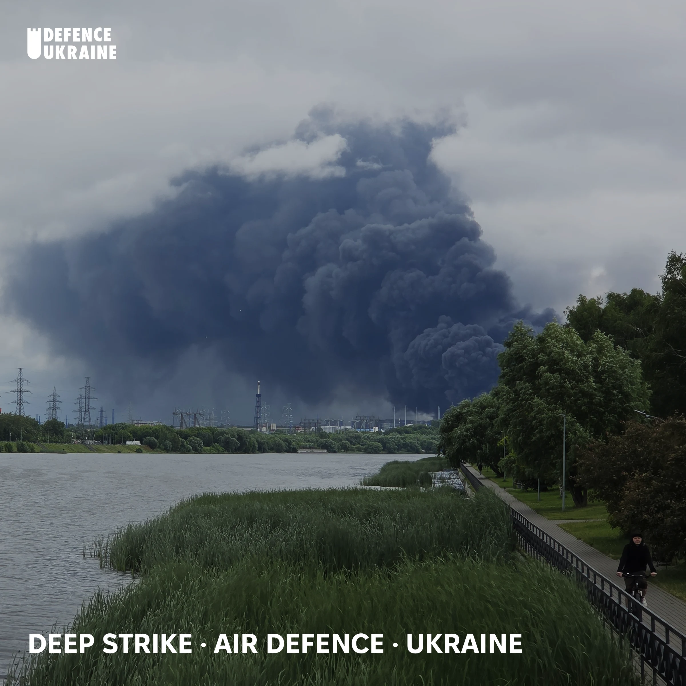 A large black smoke column rising above the Kapotnya oil refinery in southeast Moscow on 18 June 2026, seen from Brateevo Pier across the Moskva River with a cyclist passing on the embankment.