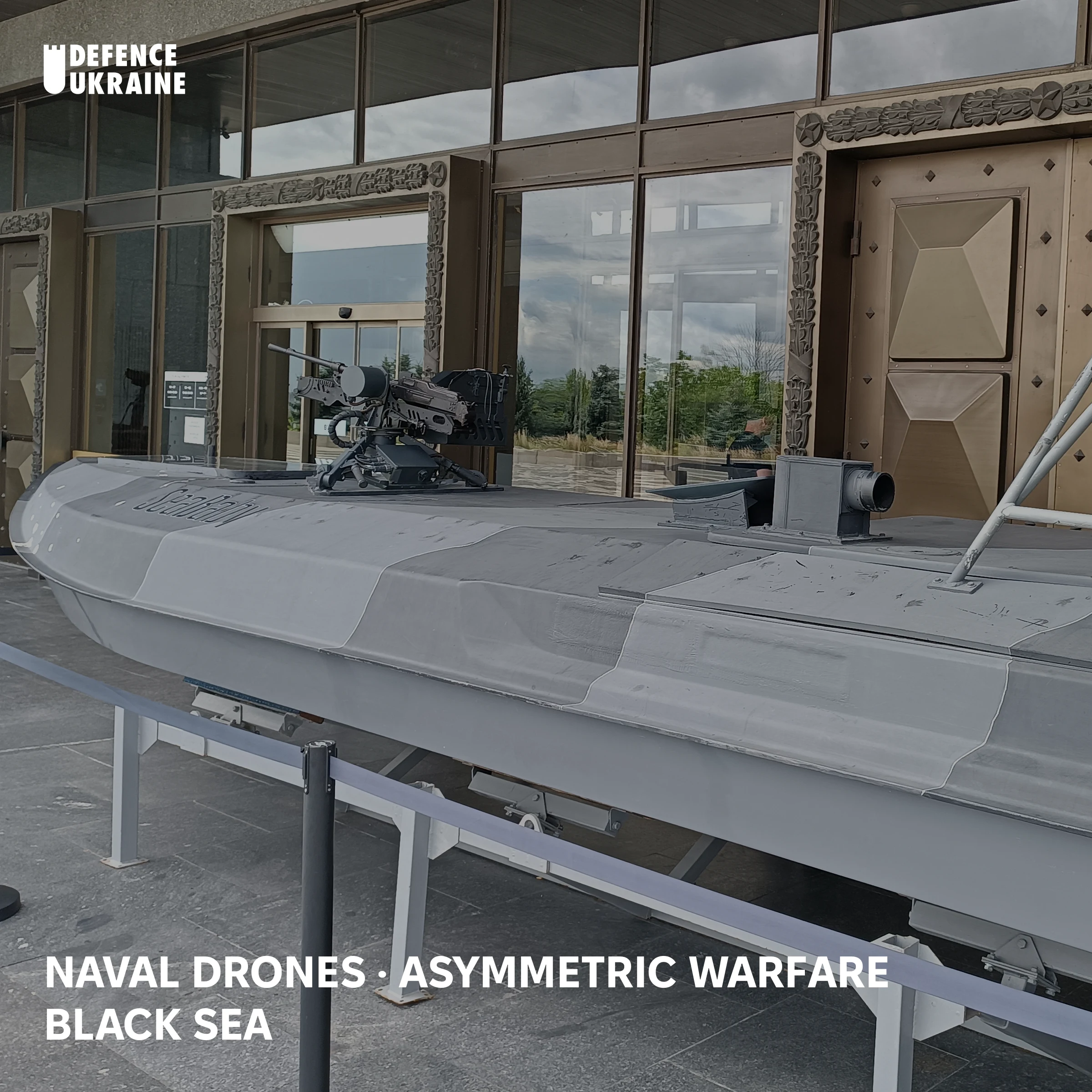 A Ukrainian "Sea Baby" maritime drone, its hull scarred from action and its Shablya remote weapon station still fitted, on public display outside the museum at Kyiv's Motherland Monument. This is the class of craft Russia's harbour booms, barges and netting are built to stop.